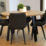Round wooden table with black chairs in a classroom with a bookshelf and plant.